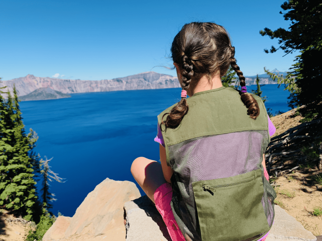 Our Junior Ranger daughter be-ing in nature at Crater Lake with deep blue waters and skies and pine trees in background