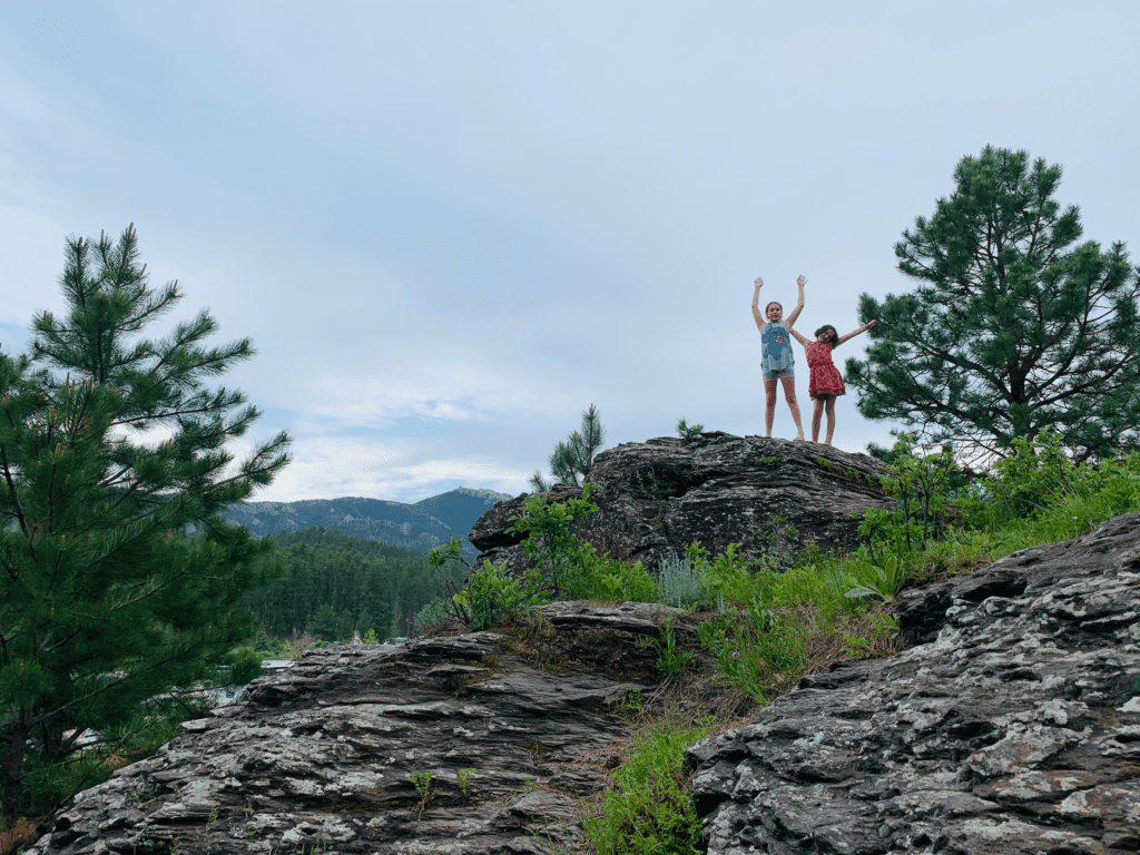 Girls climbing on rocks at the Black Hills KOA with pine trees on sides and cloudy skies in background