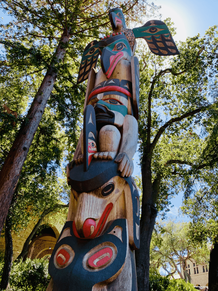 Boo-Qwilla Thunderbird Totem Pole at Stanford with thunderbird atop raven creator of earth and light and consciousness with pine trees and blue sky in background