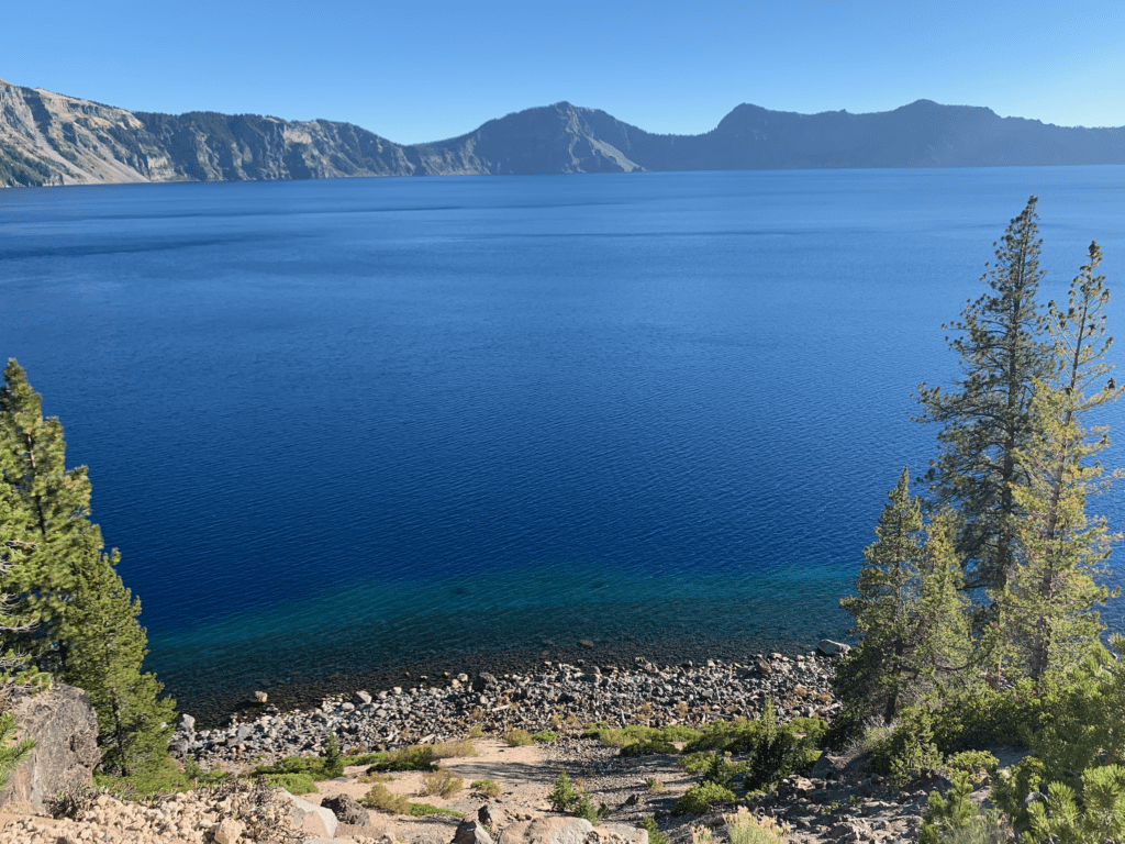 End of Cleetwood Cove Trail at Crater Lake with beach rocks and deep blue waves with conifers surround the crater rim in the background