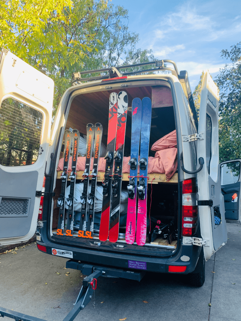 Homemade van ski rack with Dynastar SL race skis and play skis with van bed and decidious trees in the background