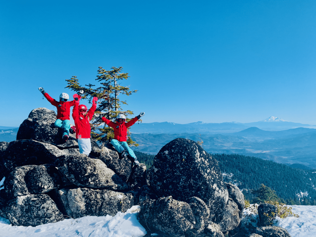 Ski kids on Mount Ashland raising their arms in the air on top of big grey boulders with a view of the Cascades and Mount Shasta in the background