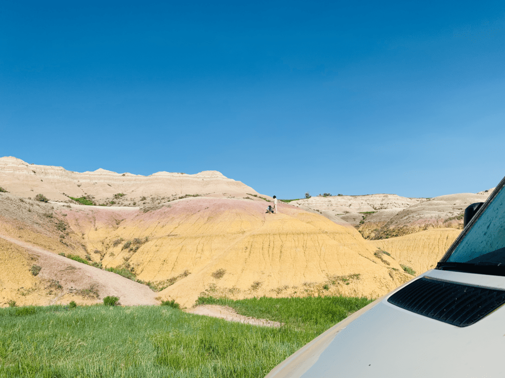 Van parked in front of rainbow rock with kids playing on top of hill with blue skies in the background at Badlands National Park