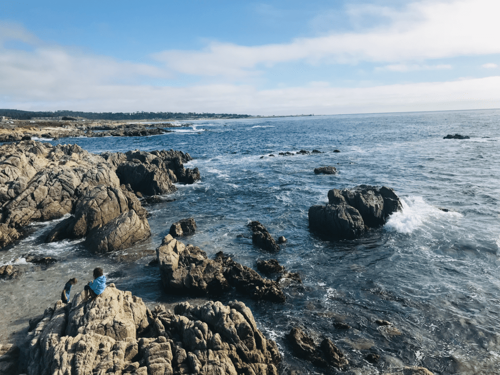 Kids sea glass hunting on the ground below at Asilomar State Beach with waves crashing against rock in the background