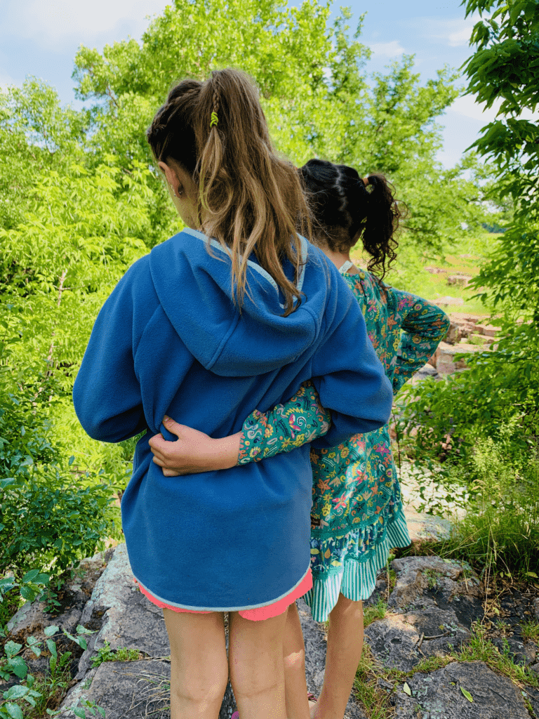 Sisters in Columbia jacket and Matilda Jane dress hugging at Pipestone National Monument with green trees in background