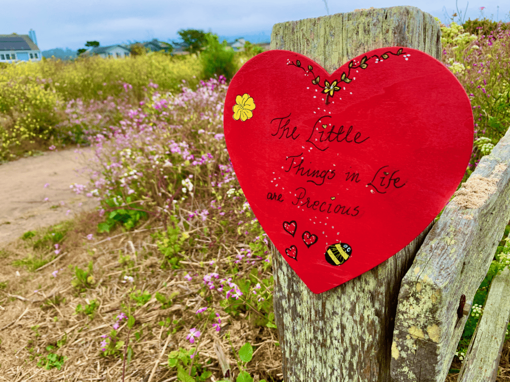 The Little Things in Life are Precious red heart sign nailed to old wooden post with yellow white and pink flower fields and blue skies in background