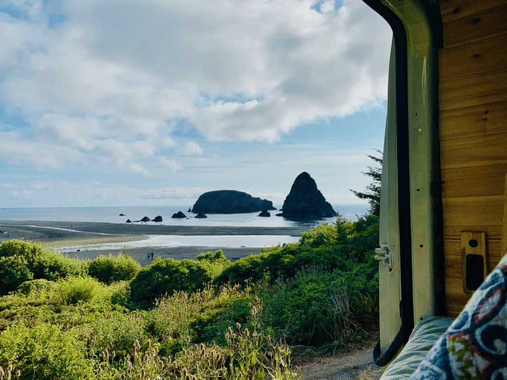 Van Life on the Oregon Coast with green shrubbery and tall dark rocks jutting out of the still blue ocean and white cumulus clouds in the background