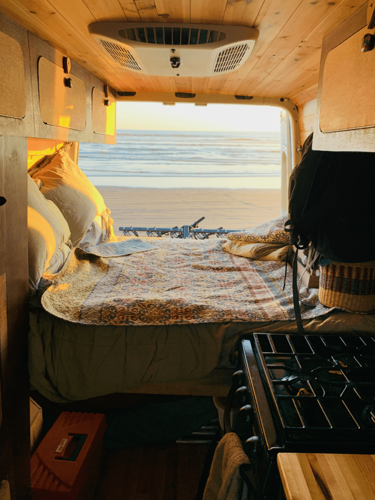 Van back doors fresh ocean air with quilt on van bed and the ocean with a sunset in the background