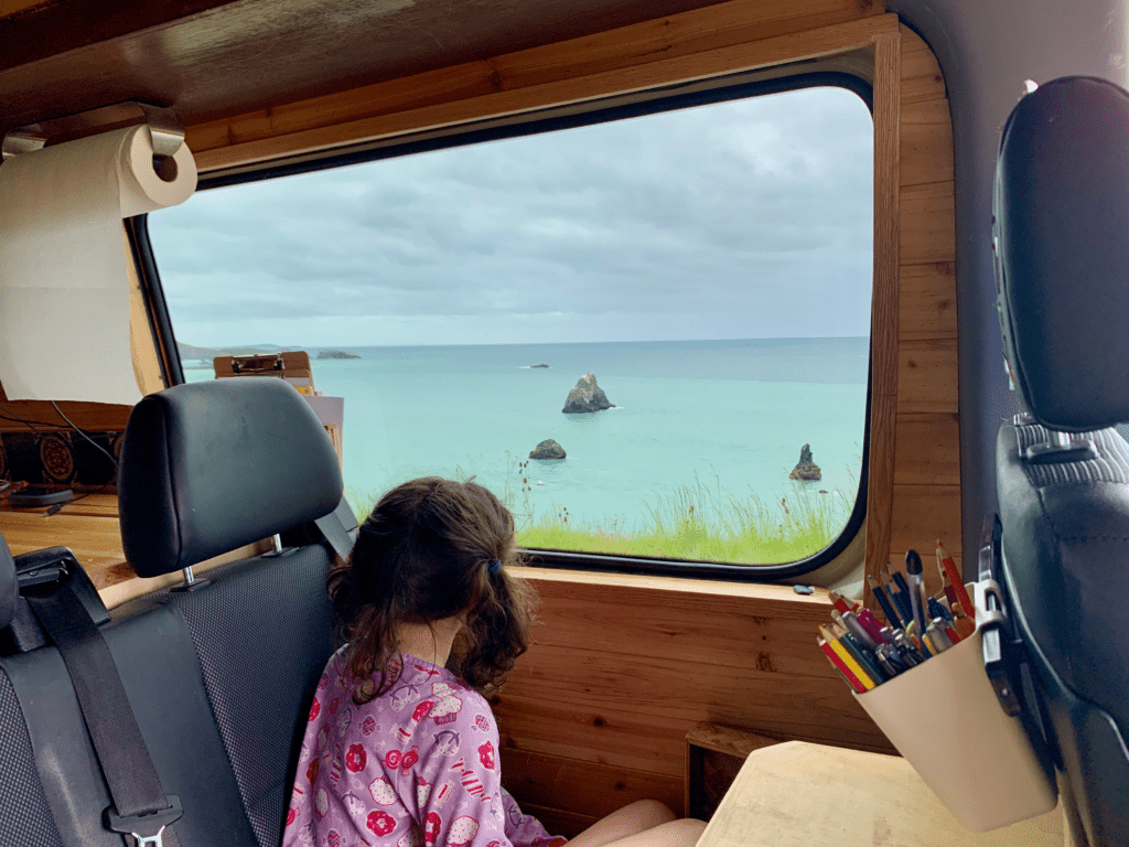 Van dining and colorful pencils art table on a ocean drive roadtrip with our daughter looking out the window at ocean rocks and waves in the background