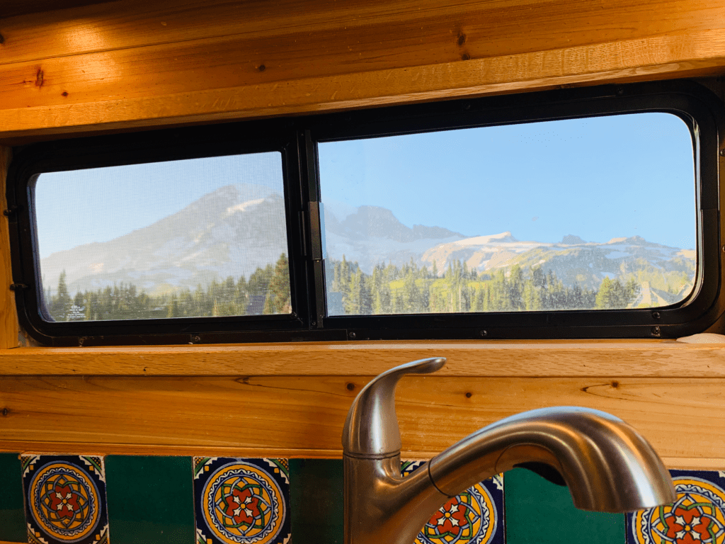 Van sliding window with mountain views over van kitchen sink with coniferous trees and mountains with blue skies in background