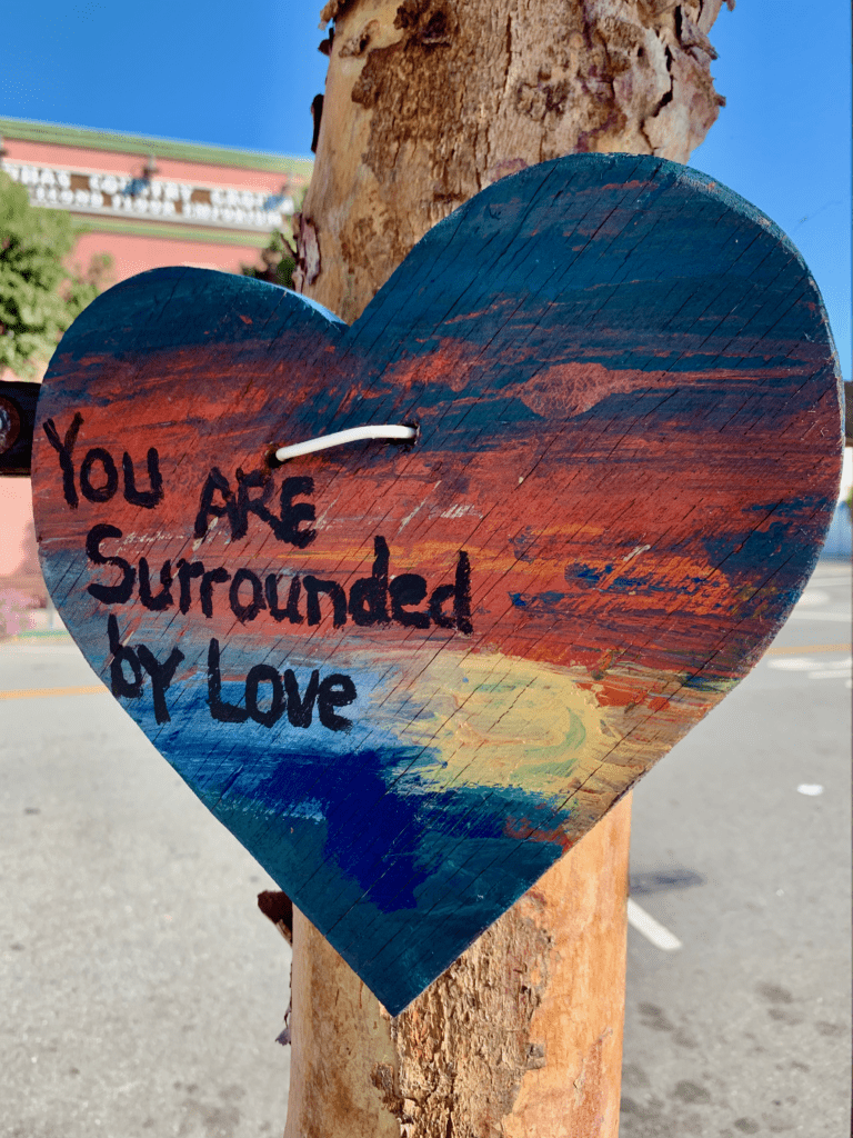 You are Surrounded by Love handprinted heart sign in Half Moon Bay California nailed to a tree with a street in the background