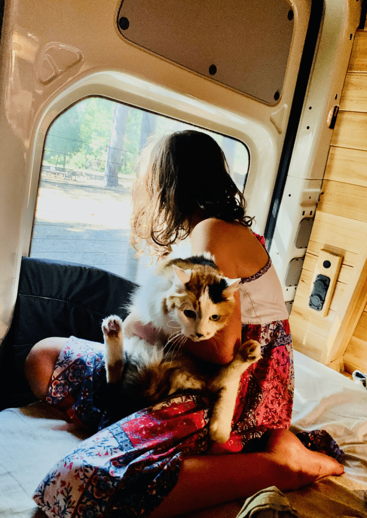 Our younger daughter holding our beloved girl Calico in the back of the van looking at the trees through the window with van build in the background