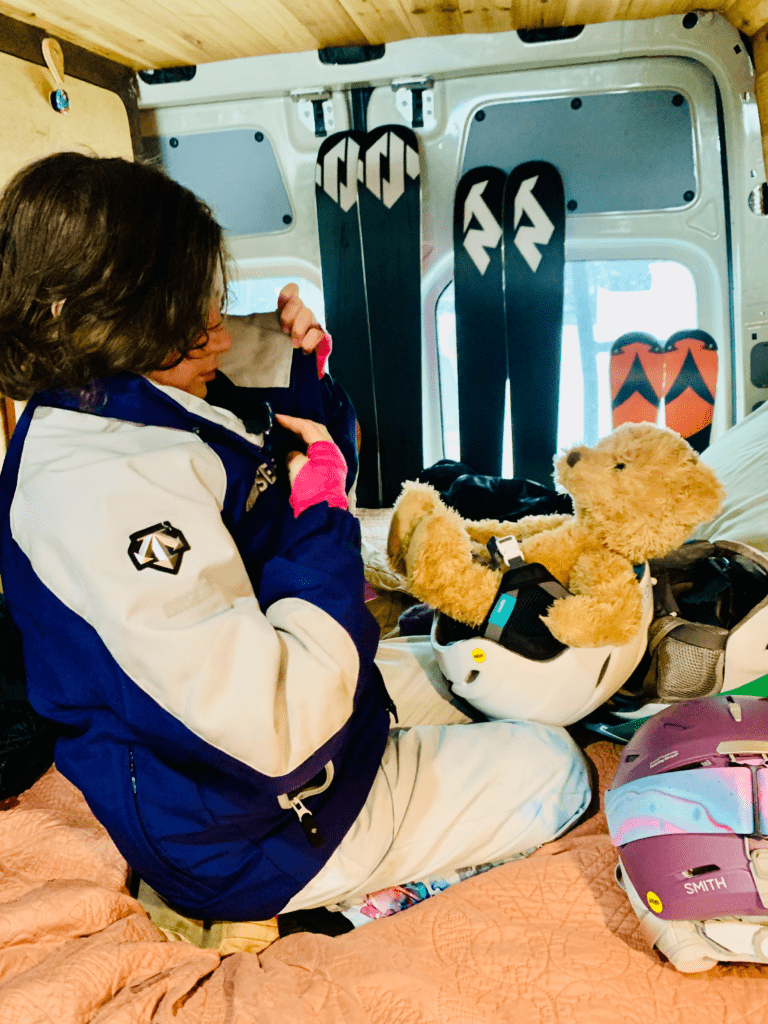 Coconut the teddy bear in a ski helmet int the Van with our younger daughter wearing a ski team jacket with ballet slippers and van ski rack in the background