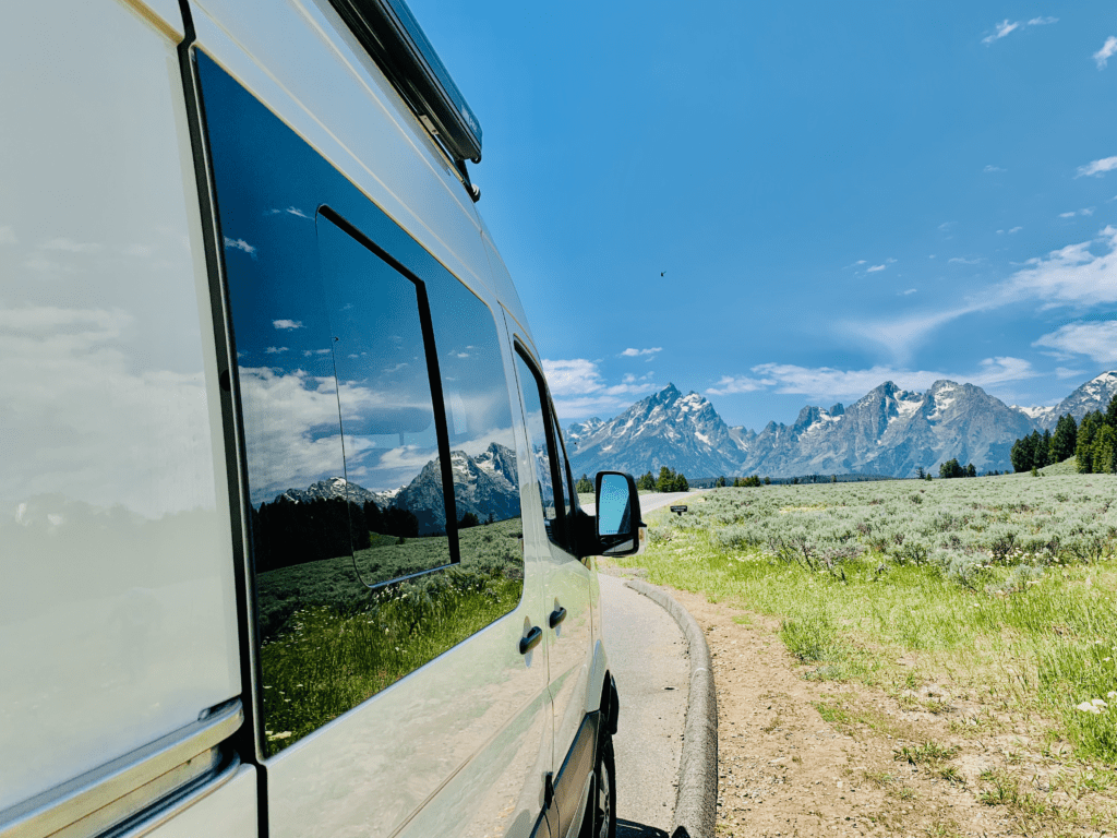 Grand Teton Van Reflection with the snow capped Tetons and wildflowers in the van window blue skies above in background