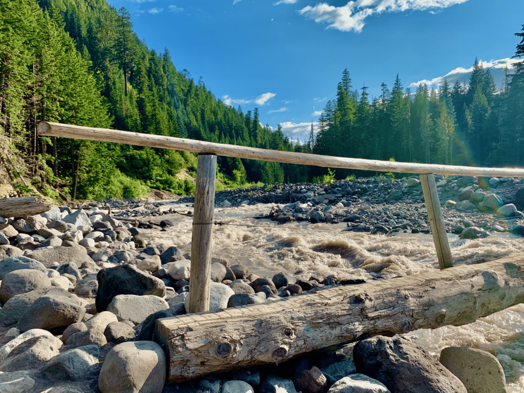 Mount Rainier wooden bridge suspended over cloudy rapid glacial silt river with blue skies and clouds in background