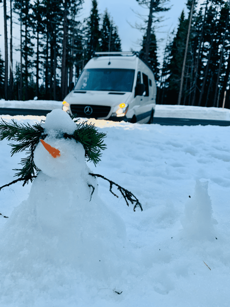 Snowman in front with orange carrot nose in Van Life ski season with Van in the background in Bend Oregon