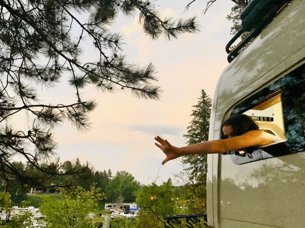 Van kid reaching Out with pine trees and an RV park with a sunset sky in the background