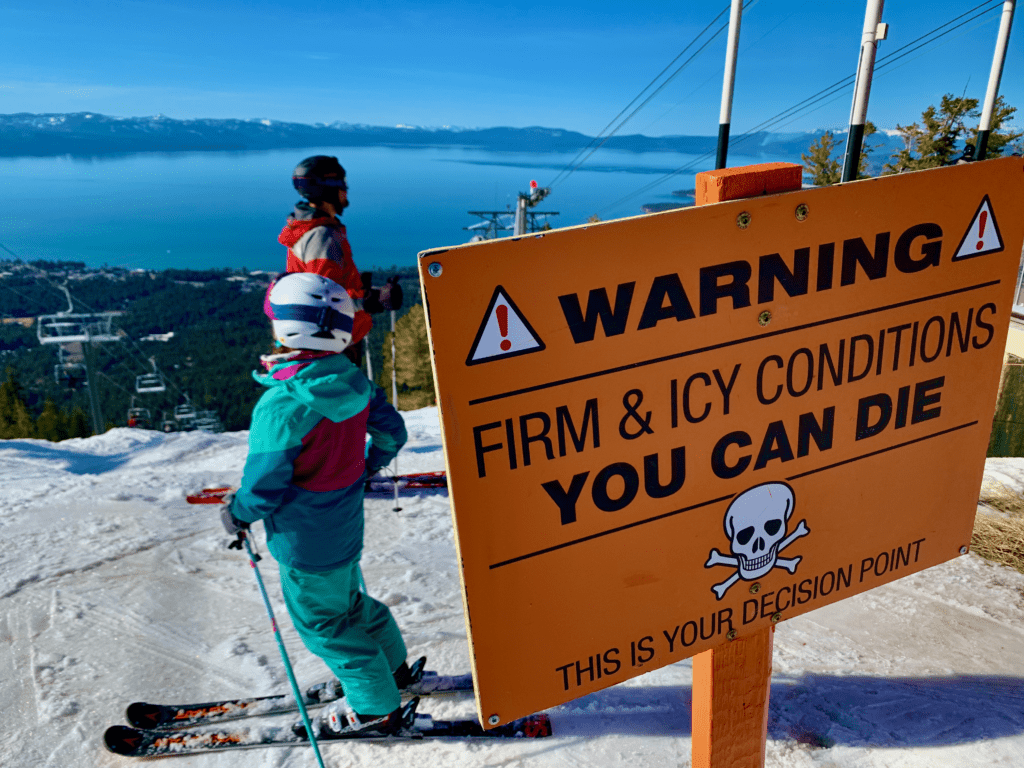 Family skiing at Heavenly with Warning Firm & Icy Conditions You Can Die Sign with Lake Tahoe in the background