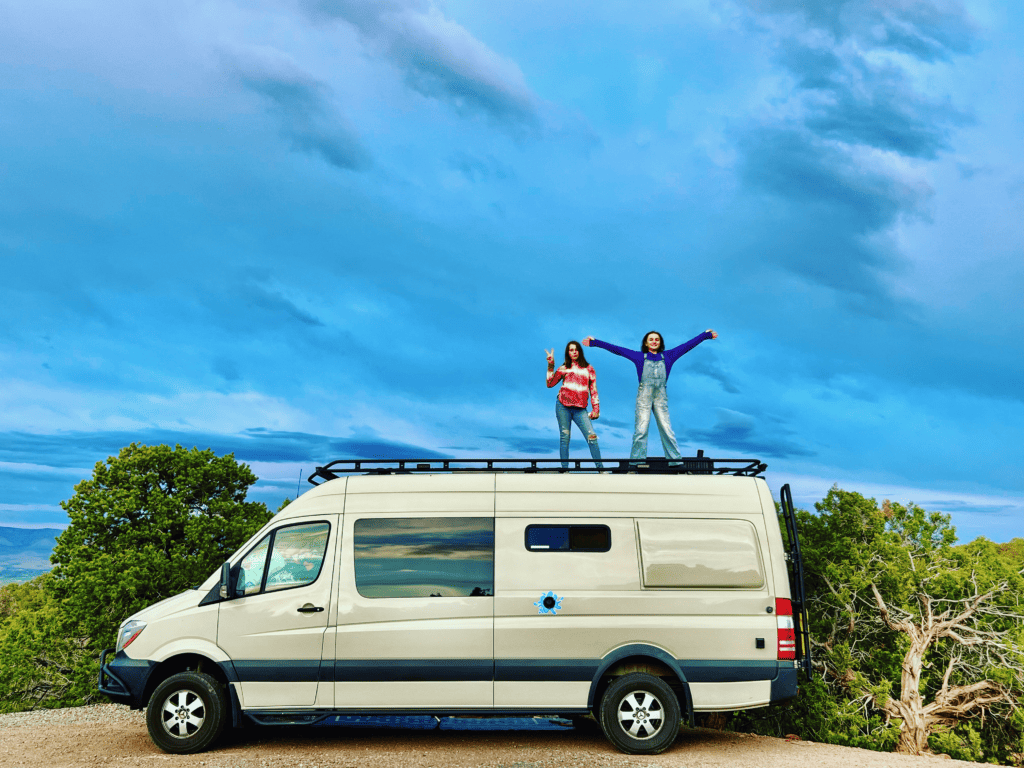 Two van kids on top of Sprinter van feeling free at Colorado National Monument with deep blue skies and Juniper and Pinyon pine trees in the background